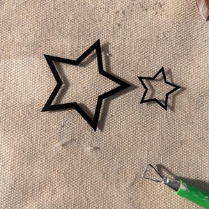 May include: Two black star-shaped stencils on a beige fabric surface. The larger star is layered on top of a smaller star.