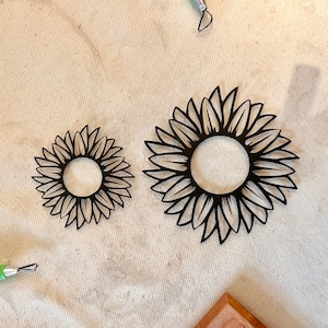 May include: Two black sunflower cutouts of different sizes. The sunflowers have a circular center and petal-like shapes around the edge. The cutouts are on a light-colored, textured surface. Crafting tools are visible in the background.