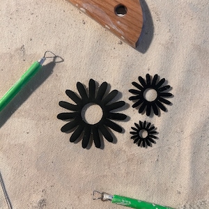 May include: Three black paper flower shapes of varying sizes on a beige fabric surface. The largest flower has a diameter of approximately 5 inches. Two smaller flowers are placed to the right of the larger flower.