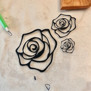 May include: Three black rose stencils on a white canvas background. The roses are different sizes and have a detailed outline.
