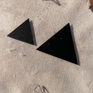 May include: Two black triangle shapes cut from paper or fabric, one larger than the other, on a beige textured surface.