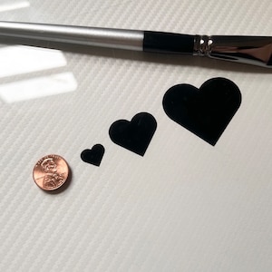 May include: Three black heart-shaped vinyl stickers of different sizes. The stickers are on a white surface with a gray pattern. A penny is included for size comparison.