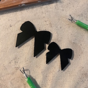 May include: Two black bow shapes made of acrylic. The bows are on a light brown fabric surface.