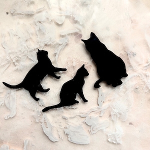 May include: Three black cat silhouettes on a white background. The cats are in different poses, one is sitting, one is standing, and one is walking.