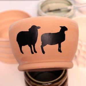 May include: A terracotta-colored ceramic bowl featuring two black sheep silhouettes. The bowl has a rounded shape with a slightly flared rim. The sheep are depicted in a simple, graphic style, adding a rustic charm to the piece.