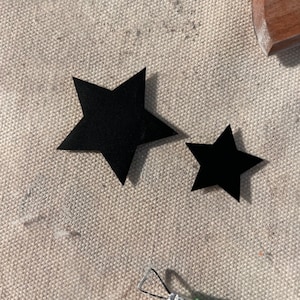May include: Two black paper stars on a beige fabric background. The larger star is on the left and the smaller star is on the right.