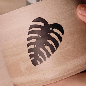 May include: A beige ceramic planter with a black silhouette of a monstera leaf.