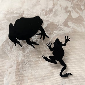 May include: Two black frog silhouettes on a white background. The larger frog is sitting with its legs spread out, while the smaller frog is standing with its legs extended.
