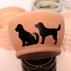 May include: A terracotta-colored ceramic pot with two black dog silhouettes. One dog is sitting, and the other is standing. The pot has a rounded shape and a slightly flared base. The background is blurred.