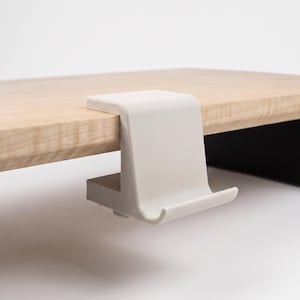 Op de afbeelding: Een matwitte, C-vormige metalen klemhaak bevestigd aan de rand van een lichtgekleurde houten plank. De haak heeft een plat bovenoppervlak dat op de plank rust en een gebogen, uitstekend onderste gedeelte ontworpen om voorwerpen op te hangen.