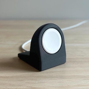 Puede incluir: Un soporte de carga negro en ángulo para un reloj inteligente. El soporte tiene una almohadilla de carga blanca y un cable blanco. El soporte está sobre una superficie de madera.