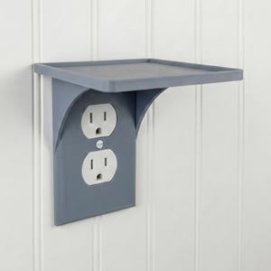 May include: A grey outlet shelf with a built-in electrical outlet. The shelf is rectangular and supported by a curved bracket. The outlet has two standard UK receptacles.