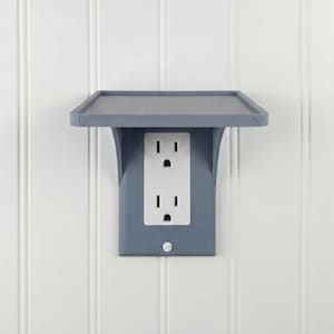 May include: A gray electrical outlet cover with a built-in shelf. The cover is mounted on a white wall with vertical paneling. The outlet has two standard three-prong receptacles.