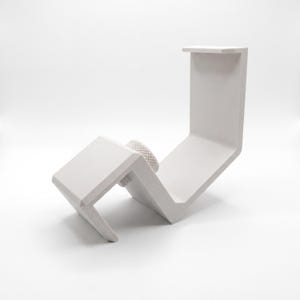 May include: A white, L-shaped clamp mount featuring a textured, matte finish. The device includes a central, circular adjustment knob with a knurled edge, designed for securing the mount to a flat surface edge with its angled, rigid structure.