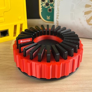 May include: A red and black game cartridge holder for Nintendo Switch games. The holder is circular with a ridged red exterior and black interior slots. Several game cartridges are visible, with the Nintendo Switch logo.