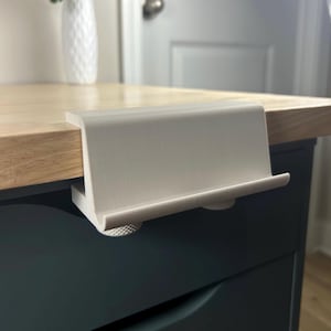Puede incluir: Un soporte para teléfono de escritorio gris claro sujeto al borde de un escritorio de madera. El soporte tiene un diseño curvo con un labio para asegurar un teléfono o tableta. La abrazadera tiene dos perillas texturizadas para apretar.