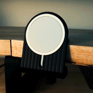 May include: A black and white MagSafe charger mounted on a wooden surface. The charger has a circular white charging pad with a black border. The mount is black with vertical grooves and a clamp to secure it to a desk or table.