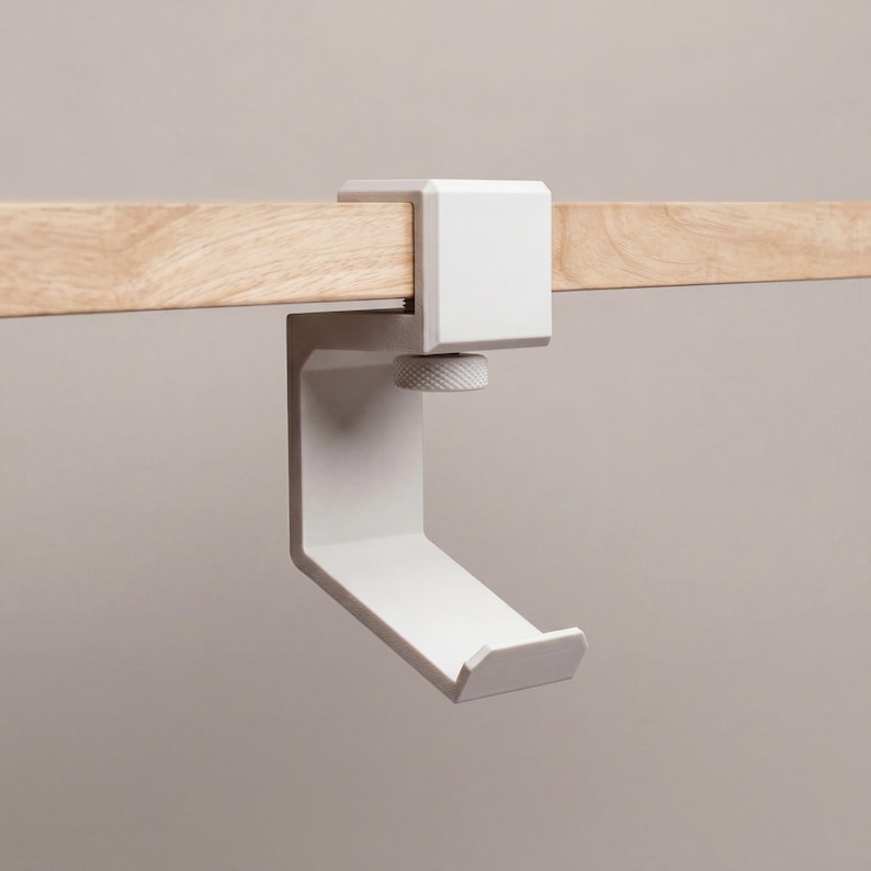 May include: A matte white C-shaped clamp hook attached to a light-colored wooden board. The hook features a textured circular tightening knob on the underside, designed to secure the bracket firmly to the edge of a flat surface or shelf.