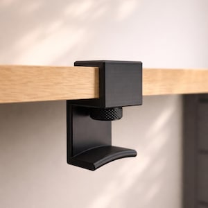 May include: A black metal clamp with a textured knob and curved support, attached to a light-coloured wooden surface. The clamp is designed to secure objects to a desk or table edge.