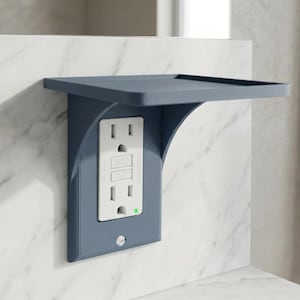 Puede incluir: Una cubierta azul para tomacorriente con un estante incorporado. La cubierta está montada en una pared de mármol blanco y presenta un tomacorriente estándar con botones "Reset" y "Test". Se ilumina una pequeña luz verde.