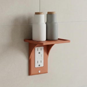 Puede incluir: Estante de pared de color terracota con enchufe eléctrico incorporado. Dos botellas cilíndricas con tapas de madera están sobre el estante. Una botella es blanca y la otra gris. El estante está montado en una pared clara.