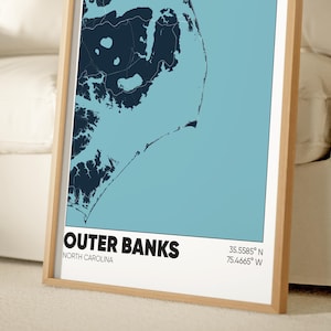 May include: A framed map of the Outer Banks, North Carolina, with a blue and white color scheme. The map shows the coastline and islands of the Outer Banks. The text "Outer Banks" and "North Carolina" is printed below the map, along with the coordinates "35.5585&deg; N, 75.4665&deg; W".