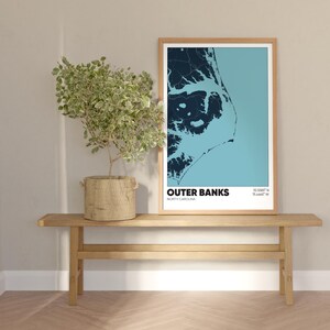 May include: A framed print of a map of the Outer Banks, North Carolina, with the text "Outer Banks, North Carolina" and coordinates "35.0088&deg; N, 75.4441&deg; W". The print is hanging on a light beige wall above a wooden bench.
