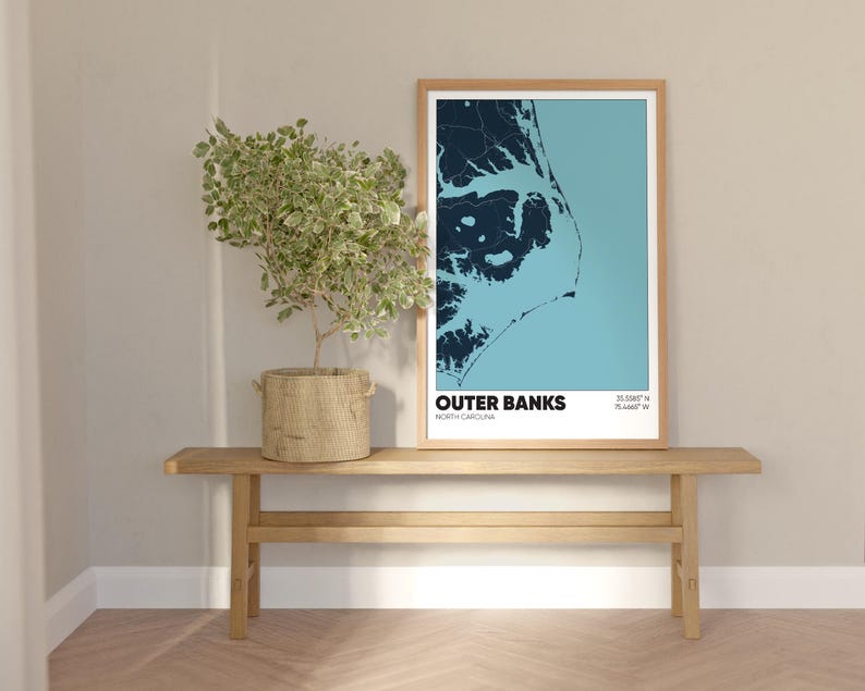 May include: A framed print of a map of the Outer Banks, North Carolina, with the text "Outer Banks, North Carolina" and coordinates "35.0088&deg; N, 75.4441&deg; W". The print is hanging on a light beige wall above a wooden bench.