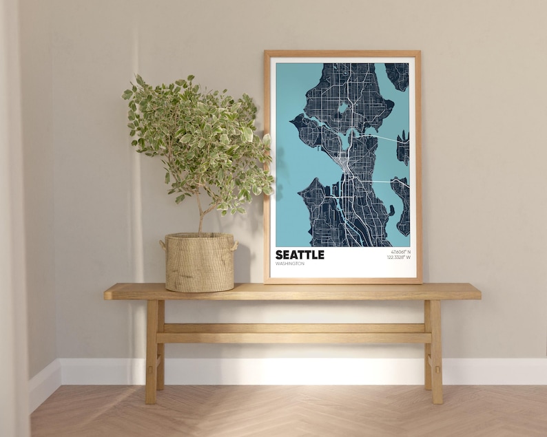 May include: A framed map of Seattle, Washington with a blue and white color scheme. The map is hanging on a light wood wall shelf with a potted plant in a woven basket.