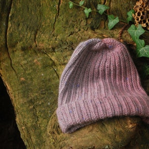 May include: A purple and grey knitted beanie hat with a ribbed texture. The hat is sitting on a tree trunk covered in moss.