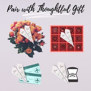 May include: A bouquet of orange roses with a paper airplane note that says "Dear From" on top of a light gray background.  There are three other images of gifts with paper airplanes on top of them: a box of chocolates, two airline tickets, and a ring in a box.