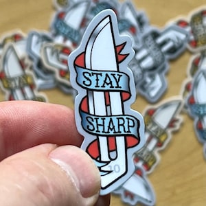 May include: A white sticker with a blue and red banner that says "Stay Sharp". The banner is wrapped around a white knife with a red handle.