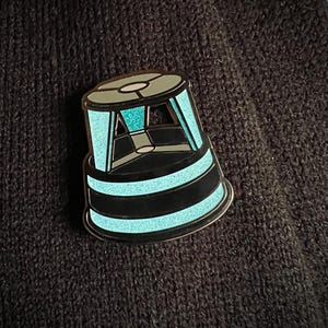 Op de afbeelding: Een teal en zwarte emaille pin in de vorm van een futuristische structuur. De pin heeft een cirkelvormige boven- en onderkant met zwarte strepen en teal glitter accenten. De pin is geplaatst tegen een donkere, getextureerde achtergrond.