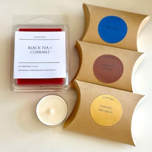 Könnte beinhalten: Eine Sammlung von duftenden Sojawachsprodukten. Enthält ein rotes Wachs-Schmelzprodukt in einem transparenten Behälter mit der Aufschrift "Black Tea + Currant", eine brennende Votivkerze und drei braune Papierverpackungen mit runden Etiketten: "Black Sea", "Hazelnut Coffee" und "Almond Macaron".