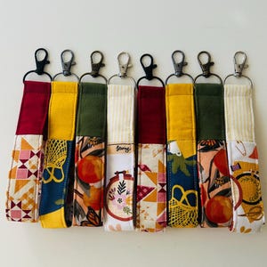 May include: Seven colorful keychains with different patterns and designs. Each keychain has a metal ring and a fabric strap with a patchwork design. The patterns include geometric shapes, floral designs, and fruit illustrations. The keychains are made of fabric and are approximately 6 inches long.