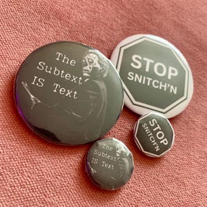 May include: Four round black and white buttons with different designs. The largest button features a black and white image of a person with the text "The Subtext IS Text". The second largest button is a stop sign with the text "STOP SNITCH'N". The two smaller buttons are identical and feature the text "STOP SNITCH'N".