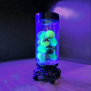 May include: A glass jar with a black lid and a decorative base sits on a wooden surface. The jar is filled with green and yellow marbles that glow under a black light.