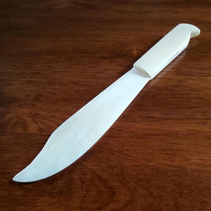 May include: A white, plastic butter knife with a rounded handle. The knife is on a wooden surface.