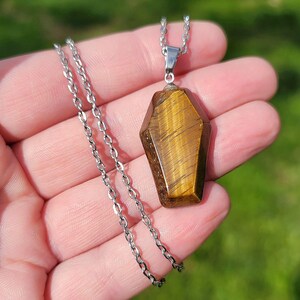 May include: A silver chain necklace with a gold tiger's eye pendant shaped like a coffin.