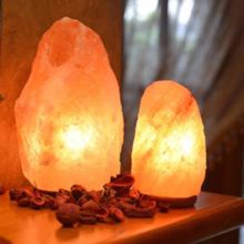 Puede incluir: Dos l&aacute;mparas de sal del Himalaya iluminadas con un c&aacute;lido brillo anaranjado. Las l&aacute;mparas tienen una textura rugosa y natural y est&aacute;n sobre una superficie de madera. Popurr&iacute; seco esparcido frente a las l&aacute;mparas.
