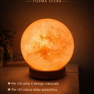 May include: A Himalayan pink salt lamp in a spherical shape. The lamp emits a warm, orange glow. The text on the image reads "Himalayan Pink Salt Lamp" and "Sphere Shape". The lamp sits on a wooden base.