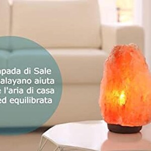 Puede incluir: Una l&aacute;mpara de sal rosa del Himalaya, que emite un brillo anaranjado c&aacute;lido. La l&aacute;mpara est&aacute; sobre una mesa blanca, con un fondo borroso. La textura rugosa y la forma natural de la l&aacute;mpara son visibles. El texto est&aacute; en italiano.