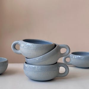 May include: A stack of four light blue ceramic cups with rounded bodies and loop handles. The cups have a textured exterior and a smooth, light blue interior. One cup is partially visible on the left, and another is on the right.