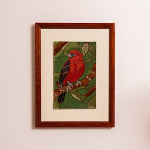 Op de afbeelding: Een ingelijst kunstwerk met een felrode vogel op een tak. De vogel heeft zwarte en rode vleugels tegen een groene en bruine achtergrond. Het kunstwerk zit in een donkerbruine houten lijst met een crèmekleurige passe-partout.