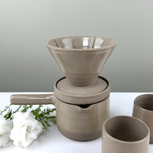 May include: A beige ceramic pour-over coffee set. It features a dripper, a carafe with a handle, a lid, and two cups. The set is designed for brewing coffee at home. The carafe has a capacity of approximately 500ml.