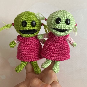 Puede incluir: Dos muñecas de ganchillo hechas a mano con cabezas verdes, ojos negros y bocas sonrientes. Llevan vestidos rosas y tienen brazos y piernas verdes. Cada muñeca tiene coletas de hilo verde claro atadas con hilo rosa.