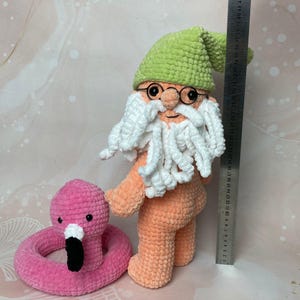 May include: A crocheted gnome doll with a green hat, white beard, and orange body. The gnome is holding a pink flamingo pool float.