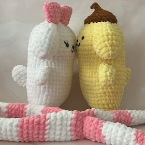 May include: Two crocheted plush toys, one white with a pink bow and the other yellow with a brown hat, are cuddling. A pink and white striped crocheted blanket is in the foreground.
