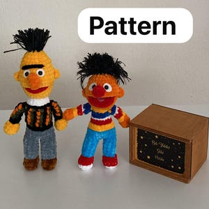 May include: Two crocheted dolls, one yellow with black hair and a striped sweater, the other orange with black hair and a striped shirt, standing next to a wooden box with the text "Bir Yıldız Gibi Parla" and a "Pattern" label above.