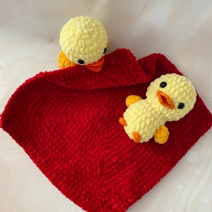 May include: A handmade crochet baby blanket in a vibrant red color, featuring two yellow duck toys. The ducks have orange beaks and black eyes. The blanket is soft and plush, perfect for a baby.